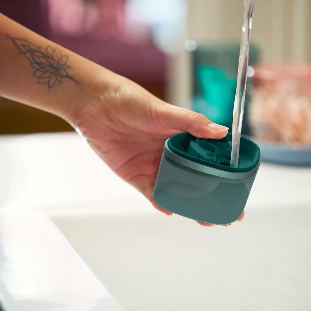 Eine Hand mit Tattoo hält einen grünen Behälter, während Wasser hineinfließt.