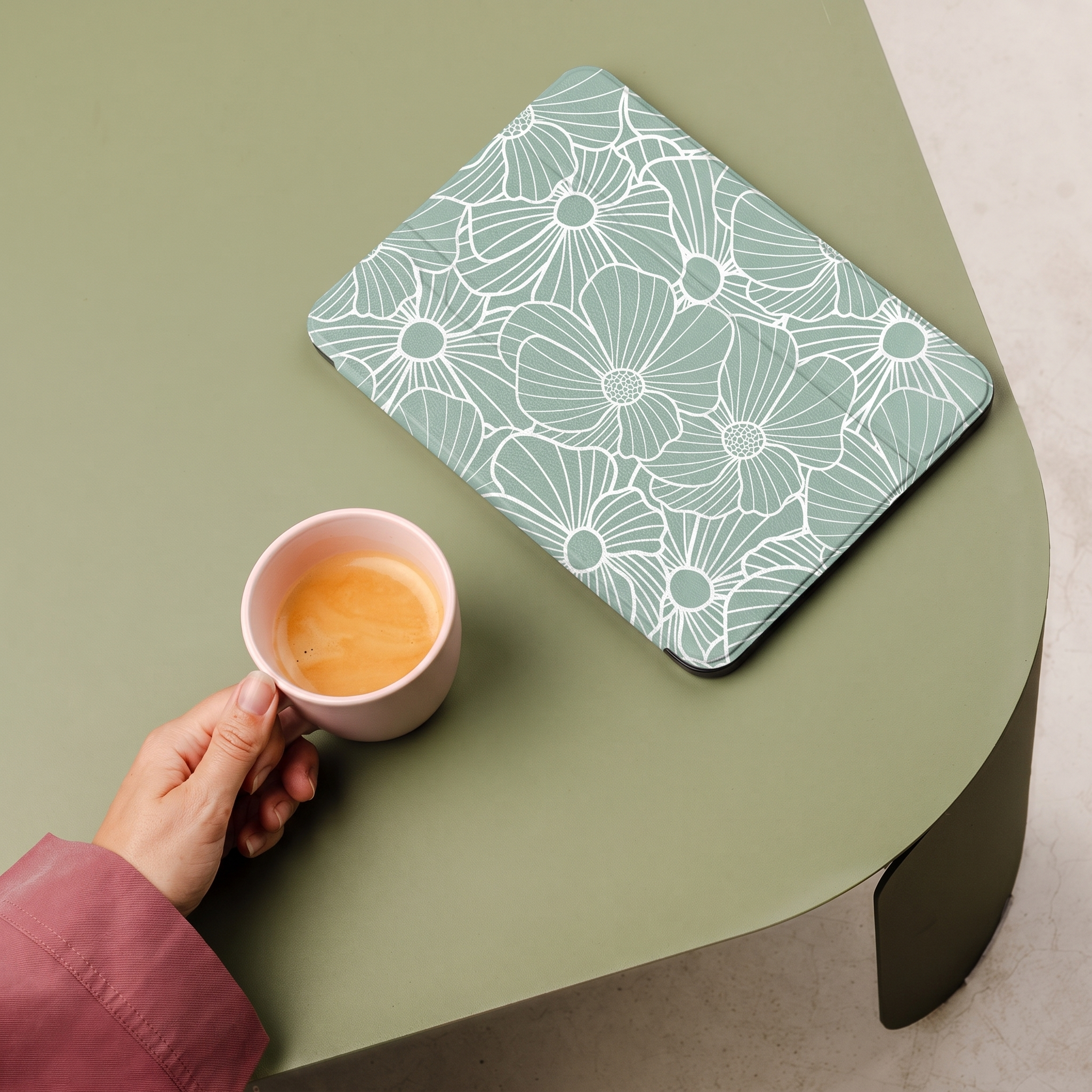 A hand holds a pink cup of coffee next to a tablet with a floral pattern.