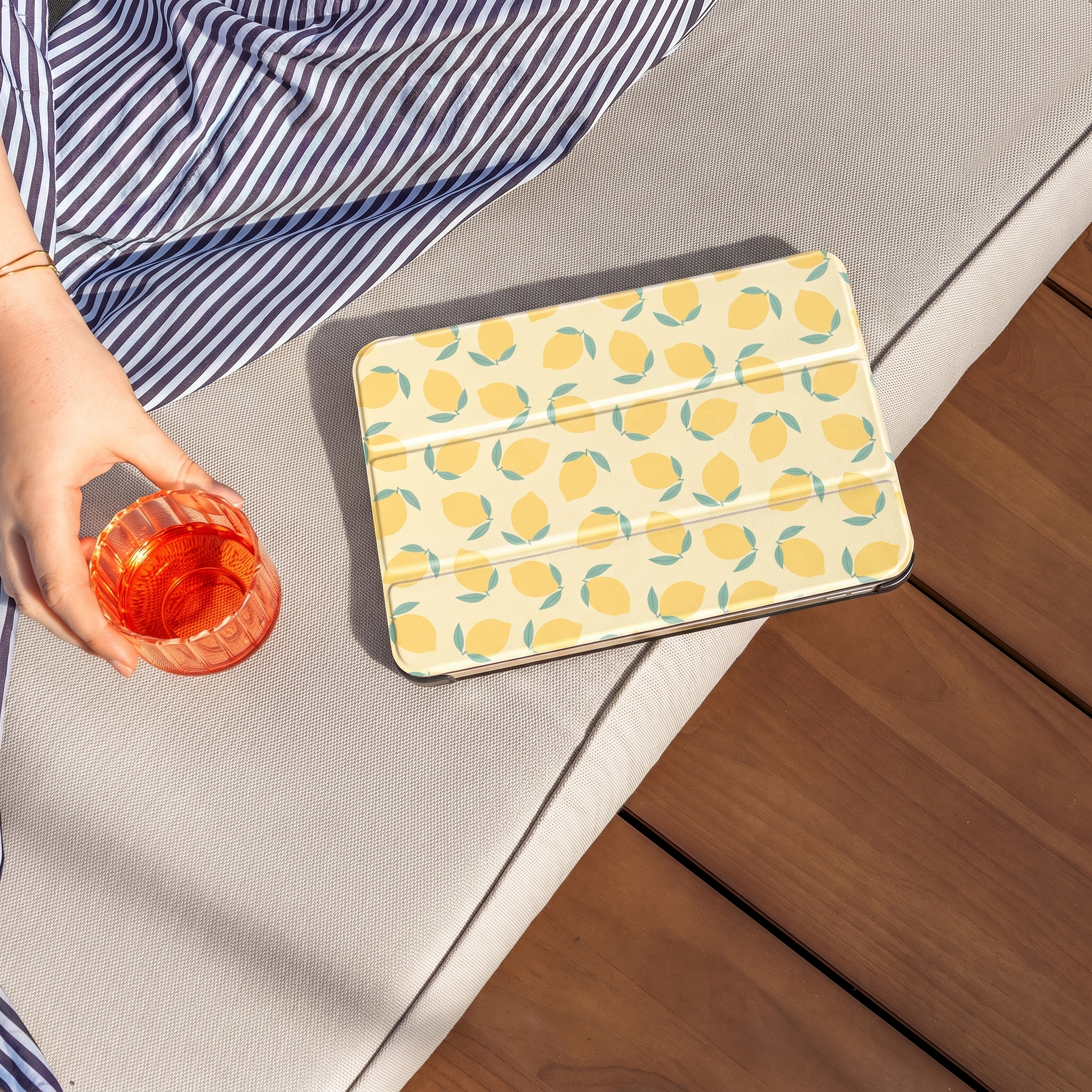 A hand holds a glass of orange drink next to a tablet with a lemon pattern.