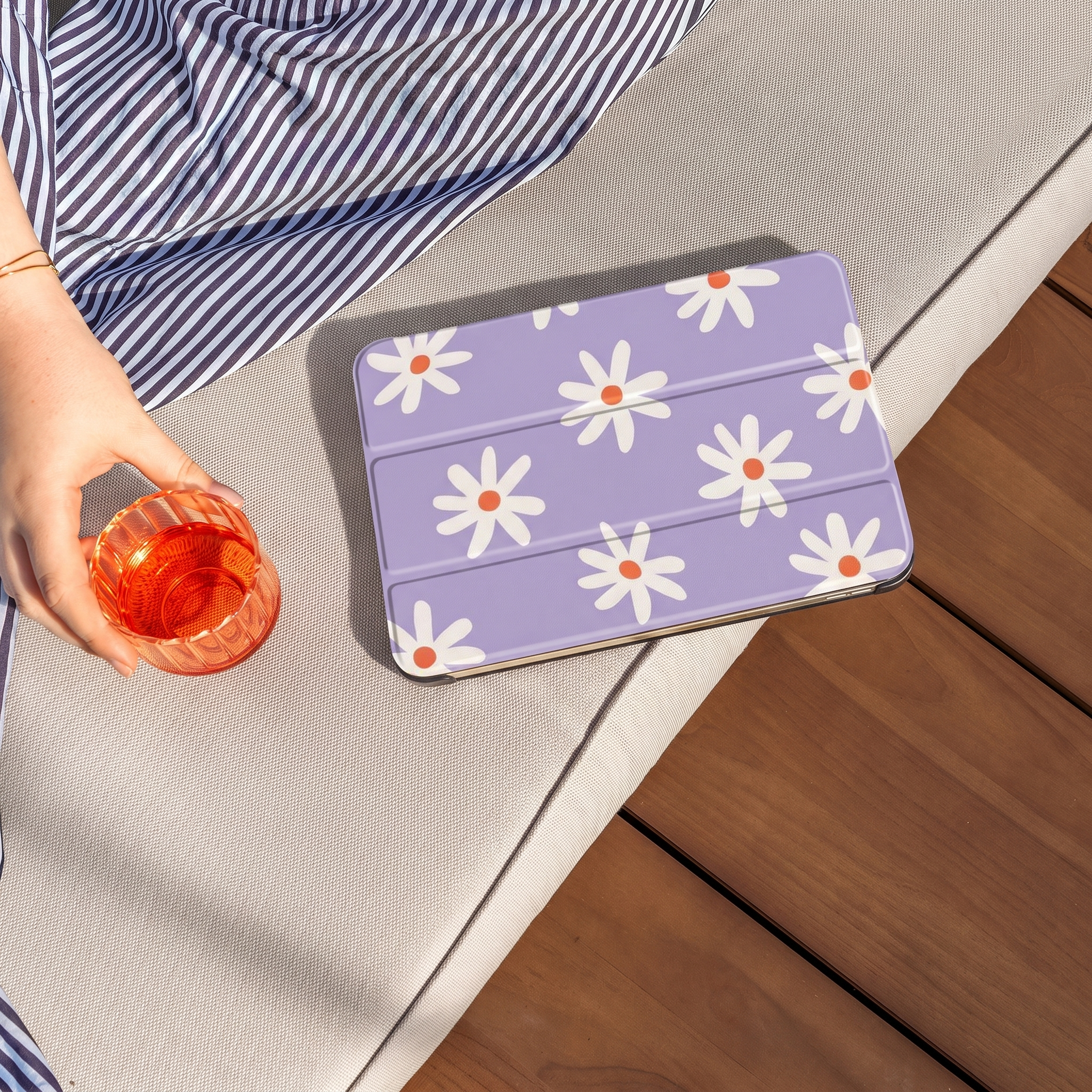 A hand holds a glass of orange drink. A purple tablet with a daisy pattern is next to it.