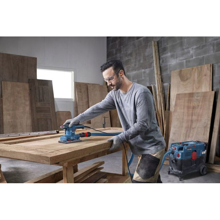 Mann schleift Holz mit blauem Schleifgerät, verbunden mit Staubsauger.