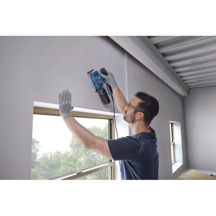 Mann schleift Wand am Fenster mit blauer Schleifmaschine. Trägt Handschuhe und Brille.