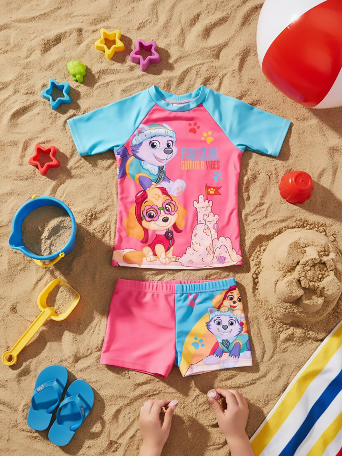 Paw Patrol Badeanzug und Shorts auf Sand mit Strandspielzeug und einem Ball.