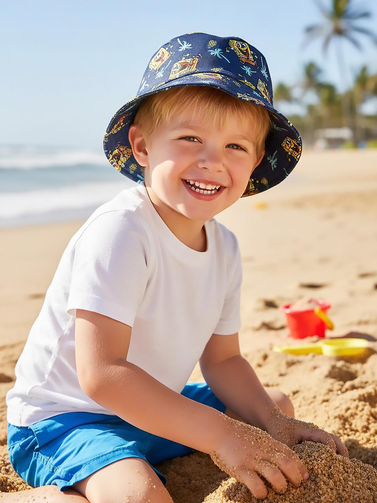 Ein lächelnder Junge mit Spongebob-Hut spielt am Strand mit Sand. Blaue Hose, weißes Hemd.