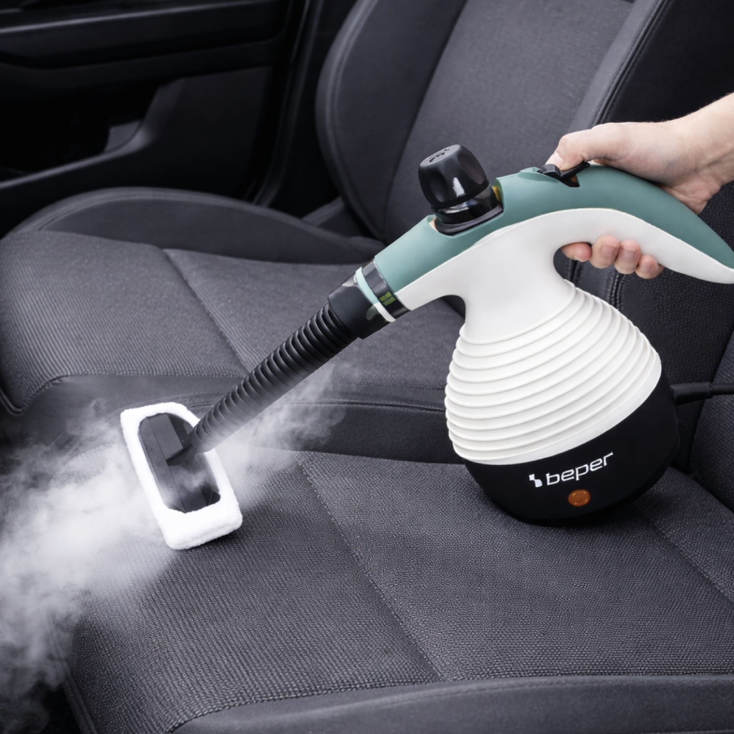 Una mano tiene un pulitore a vapore, pulendo un sedile auto grigio scuro. Il vapore è visibile.