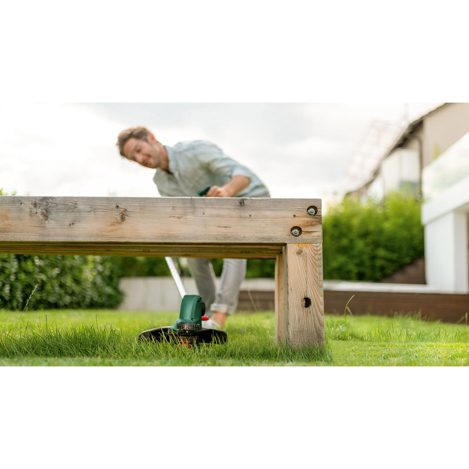 Mann trimmt Gras neben Holzstruktur mit elektrischem Trimmer im Garten.