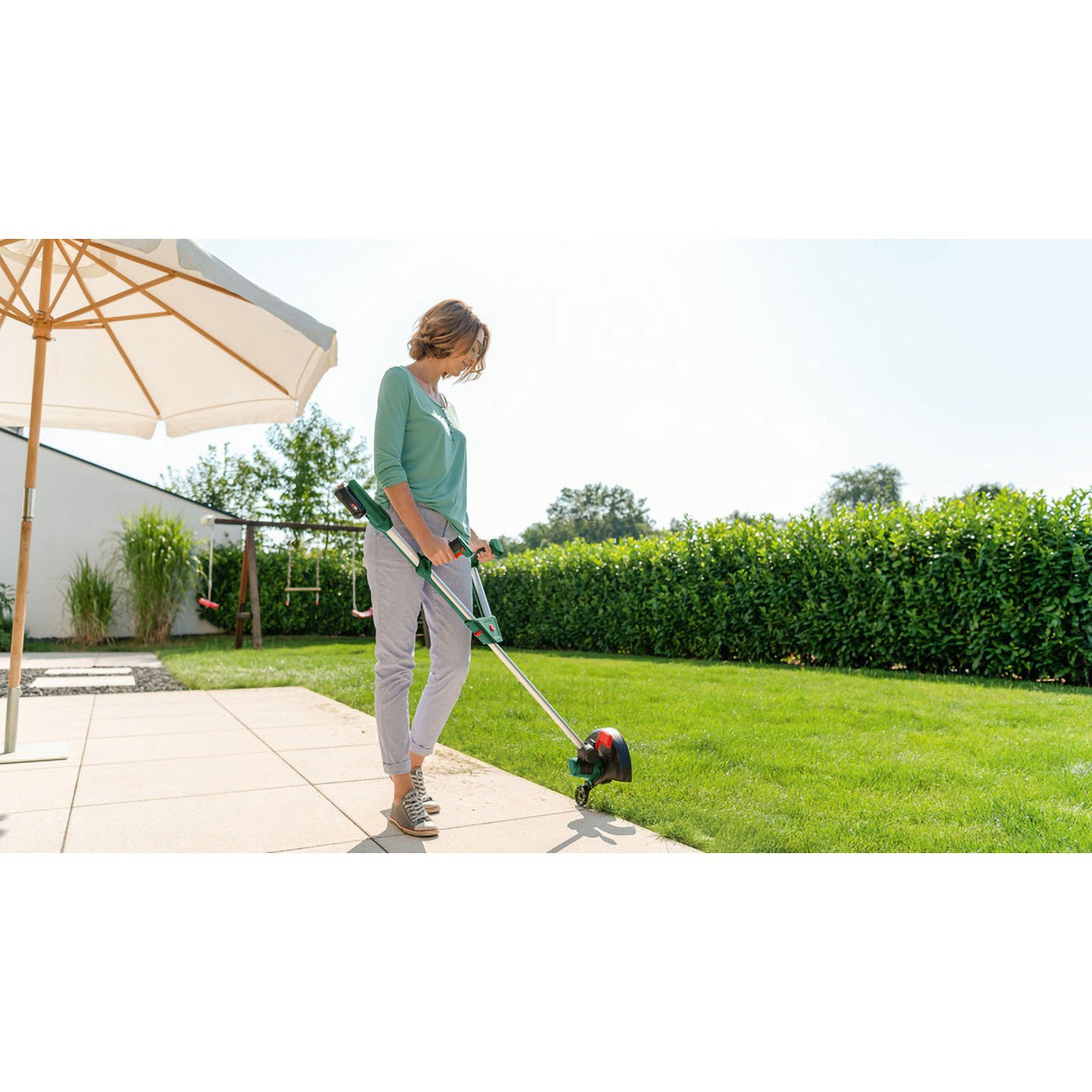 Frau trimmt Rasen im Garten mit einem Rasentrimmer neben Terrassenplatten.