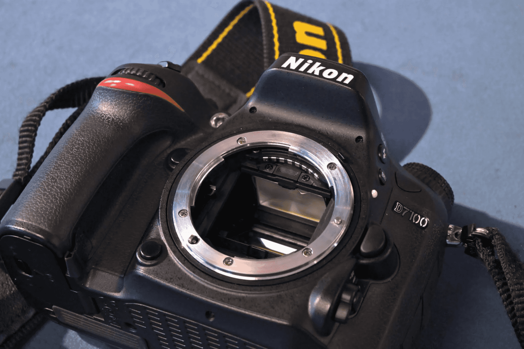 Fotocamera reflex Nikon D7100 nera con attacco obiettivo aperto, tracolla nera con finiture gialle.