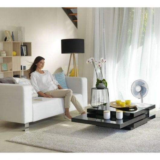 Una mujer se sienta en un sofá blanco en una sala de estar luminosa con un ventilador y una mesa.