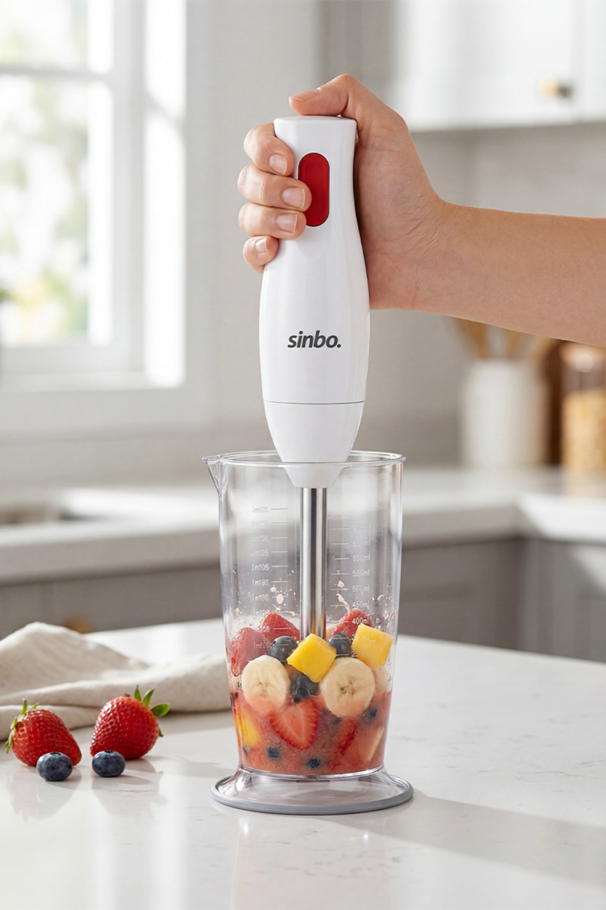 Çilek, yaban mersini, muz ve mango meyveleri olan bir fincanın üzerinde beyaz Sinbo blender tutan el.