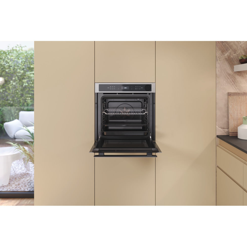 Un forno nero con lo sportello aperto è integrato in un mobile da cucina beige, con un giardino verde visibile fuori da una finestra.