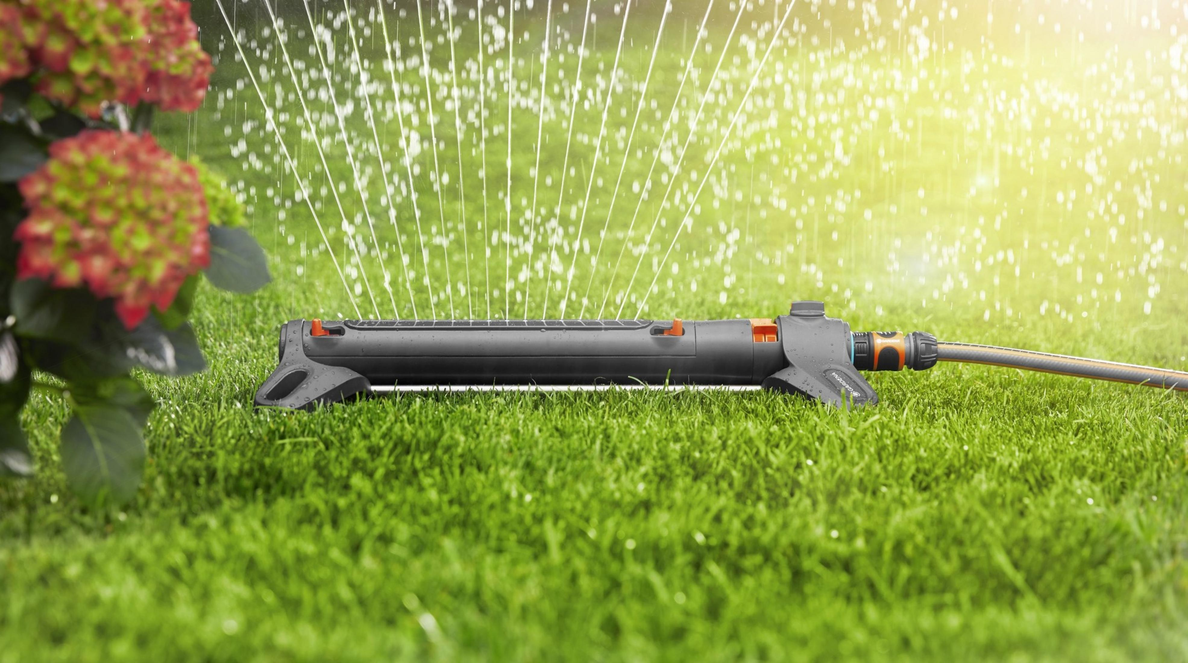 Un irrigatore da prato innaffia l'erba con fiori rossi e verdi sullo sfondo.