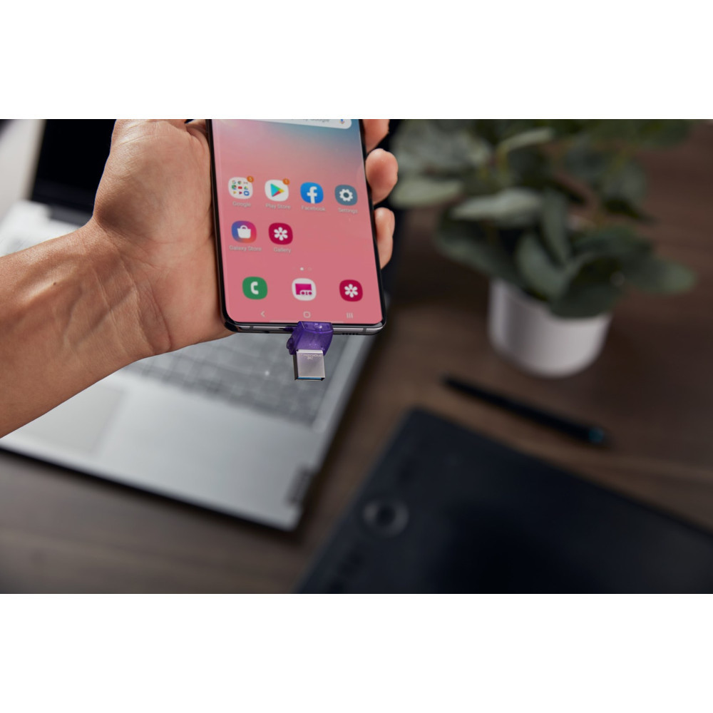 Una mano tiene uno smartphone con un'unità USB. Un laptop e una pianta sono sullo sfondo.