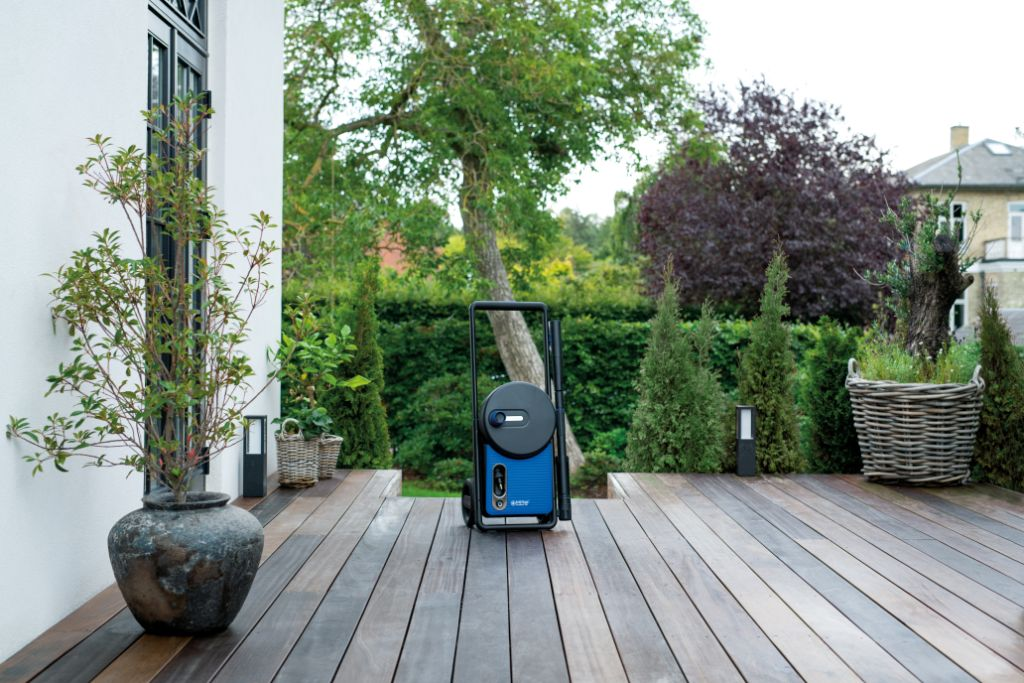 Ein blauer Hochdruckreiniger steht auf einer Holzterrasse neben Pflanzen.