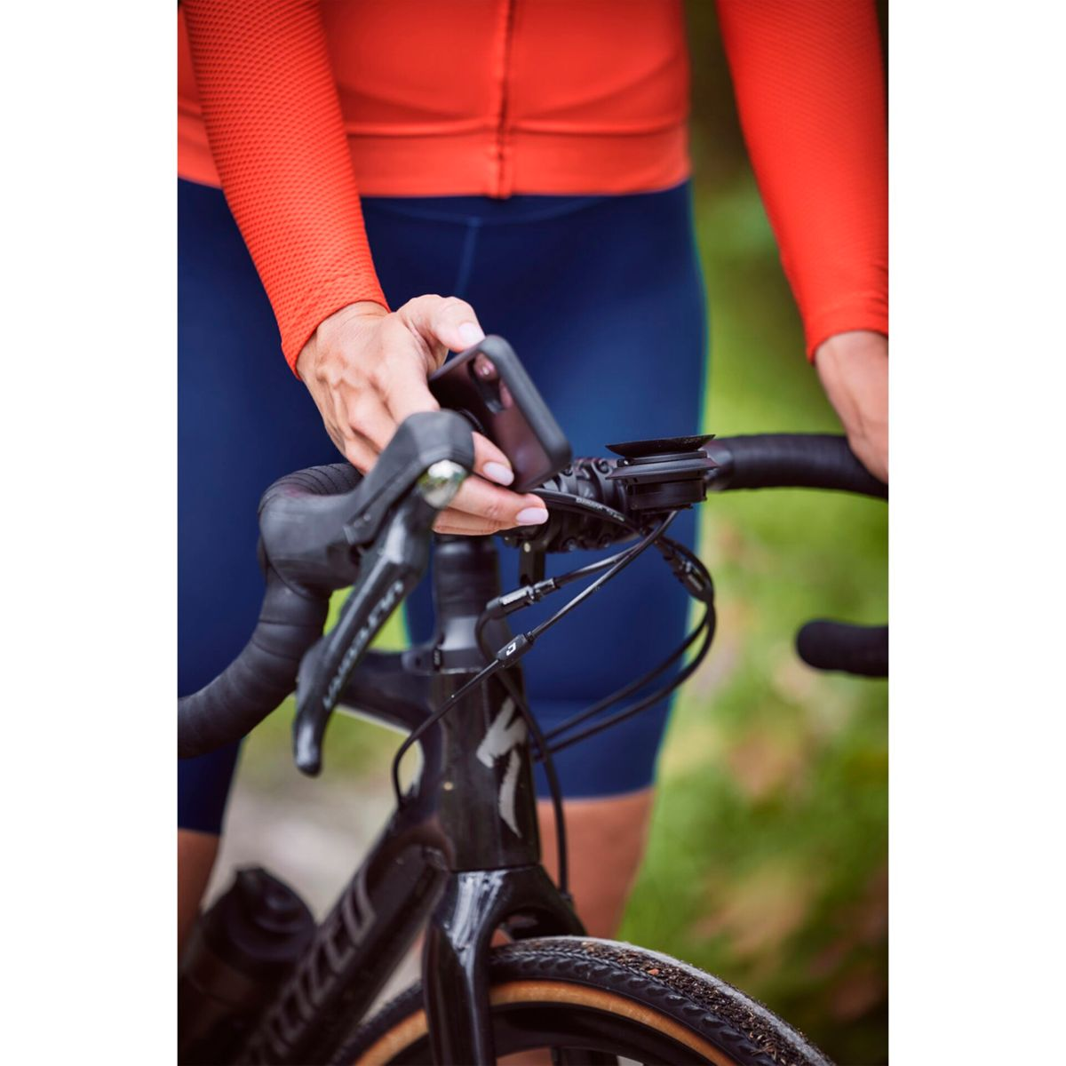 Ciclista tiene il telefono sul manubrio. Giacca arancione, pantaloncini blu.