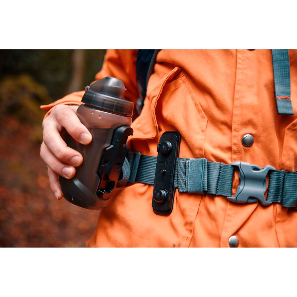 Eine Hand hält eine braune Wasserflasche vor einer orangefarbenen Jacke und einem Gürtel.