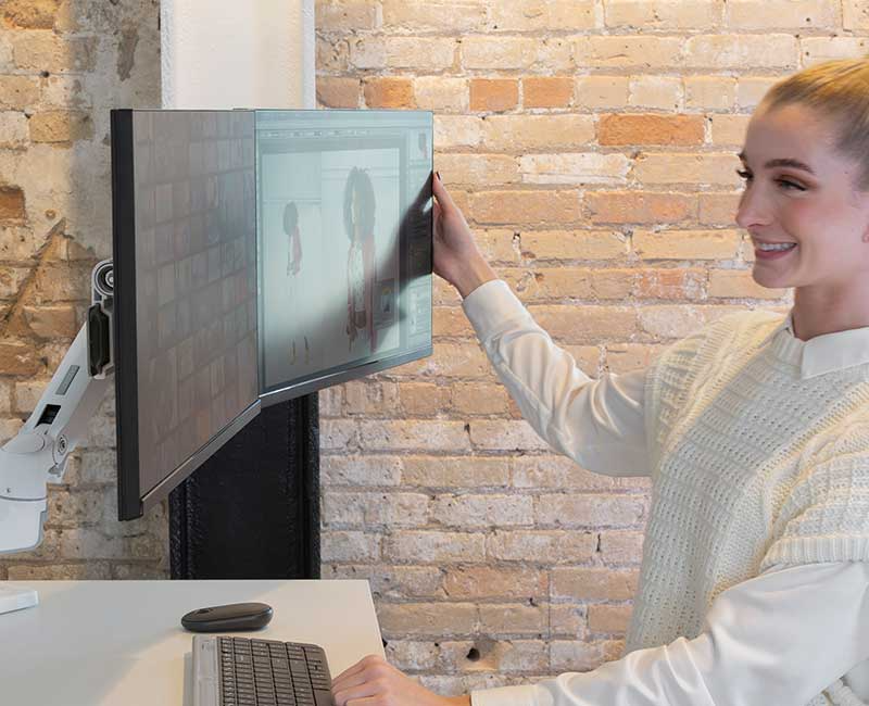 Kobieta reguluje monitor na biurku. Nosi biały sweter kamizelkę.