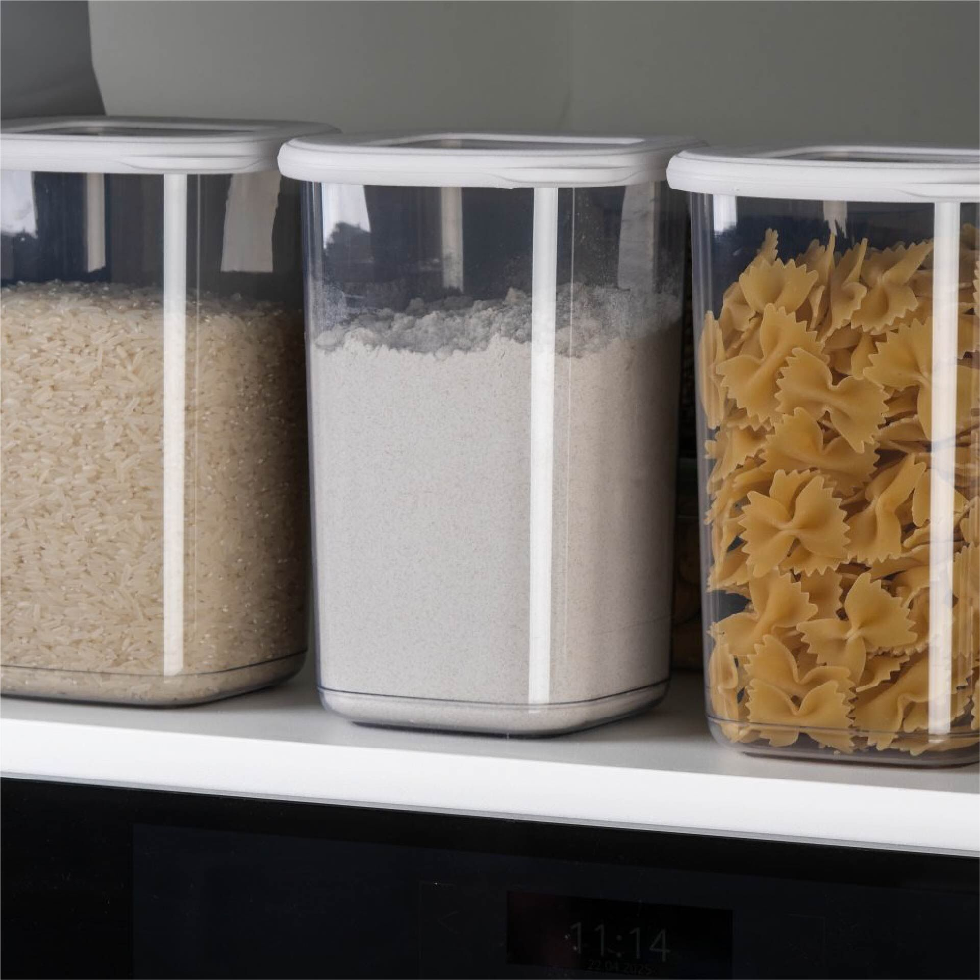 Trois récipients de stockage transparents sur une étagère. Gauche : riz. Milieu : farine. Droite : pâtes farfalle.