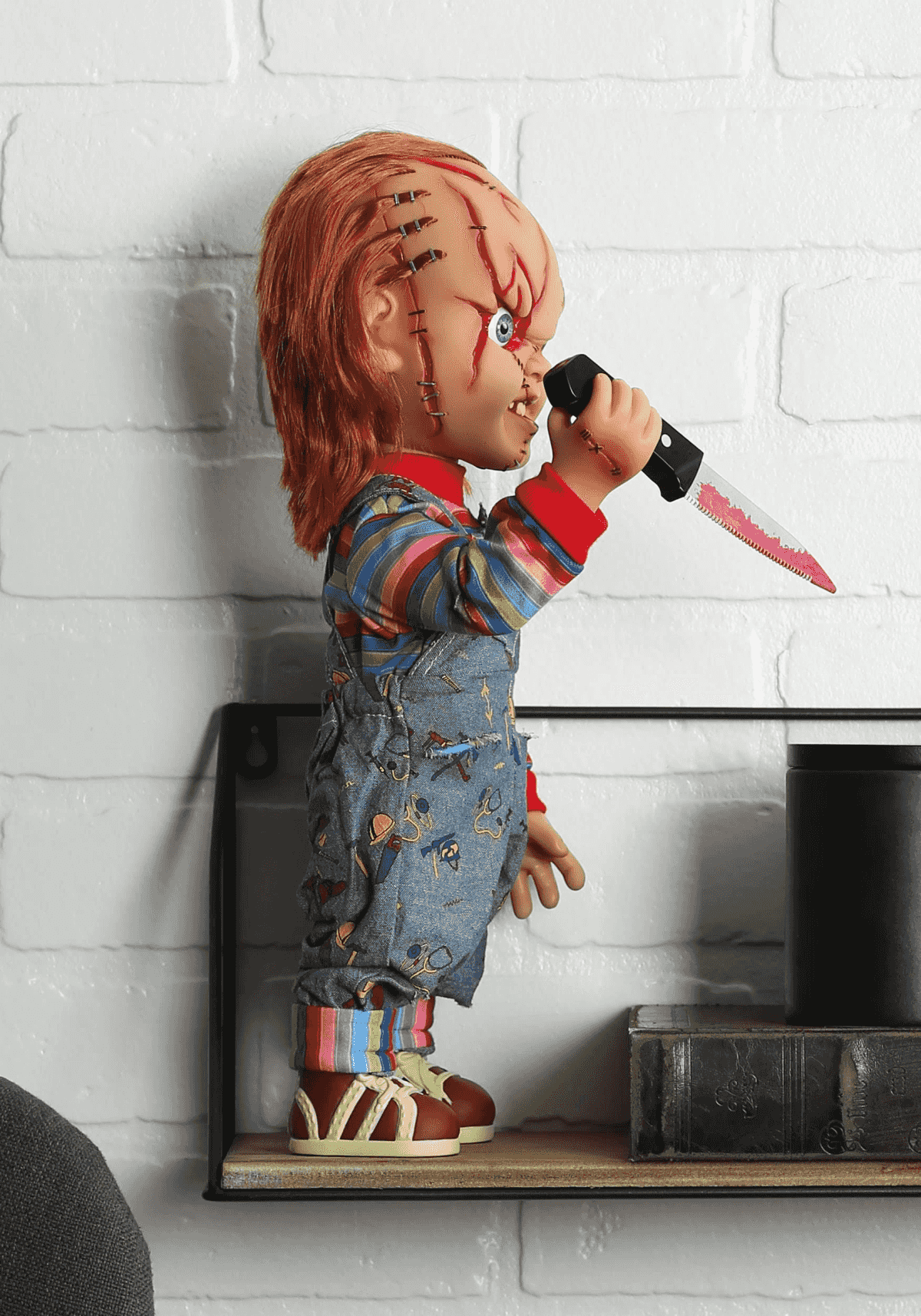 Muñeco Chucky con cuchillo, de pie en una estantería contra pared de ladrillo blanco.