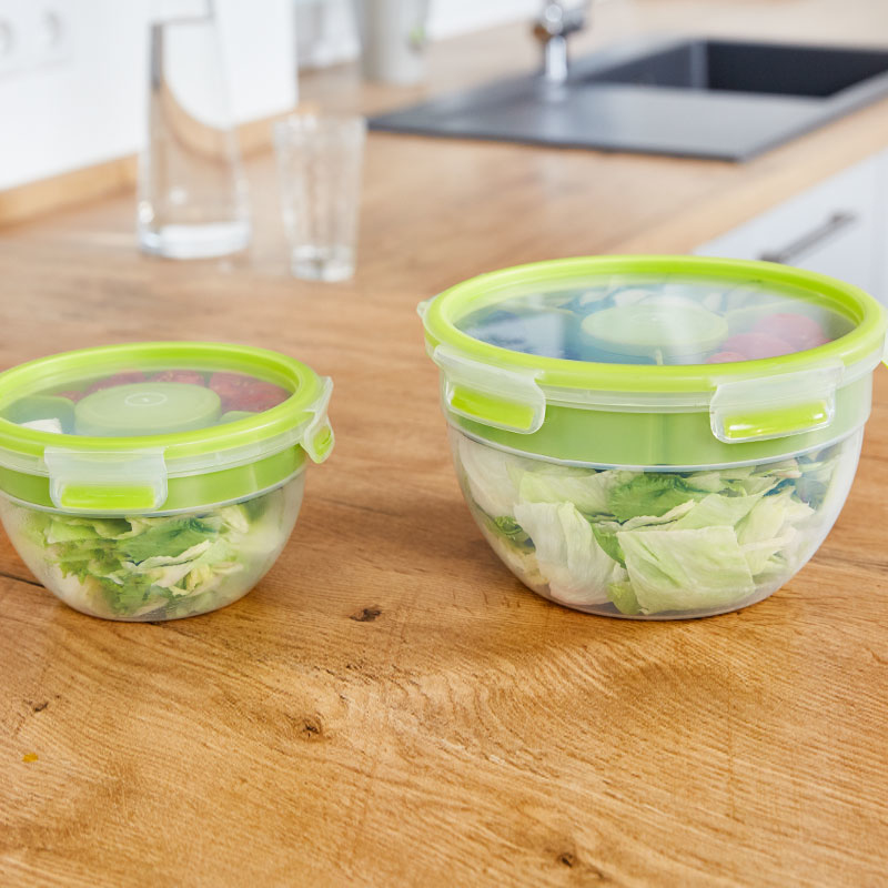 Zwei grüne Frischhaltedosen mit Salat und anderen Zutaten auf Holztisch.