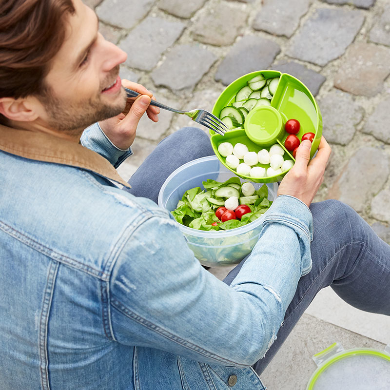 Mann im Jeansjacke isst Salat aus grünem Behälter mit Fächern. Enthält Gurken, Tomaten und Mozzarellakugeln.