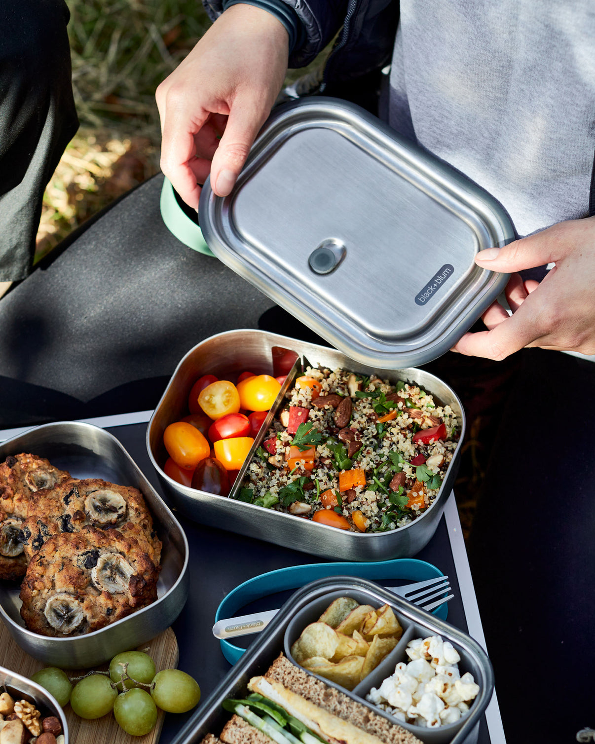 Hände öffnen eine Lunchbox mit Quinoa-Salat, Kirschtomaten, Keksen und Sandwiches.