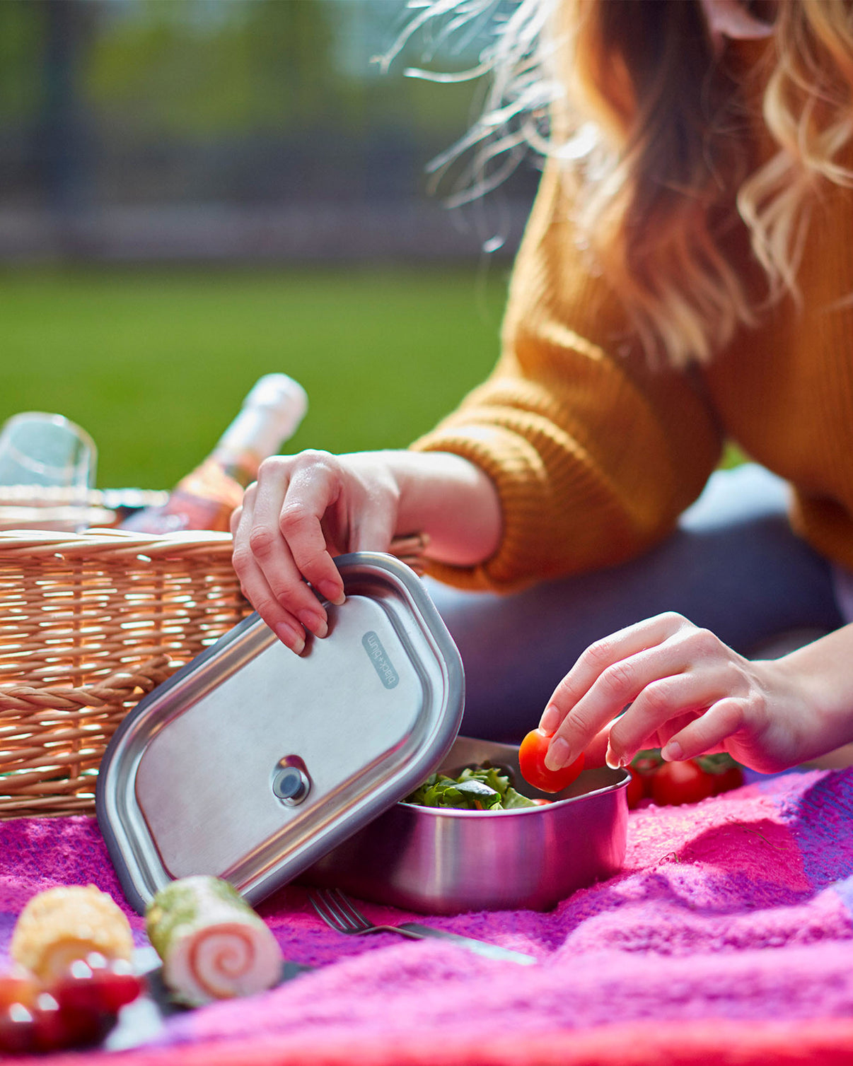 Frau packt einen Picknickkorb aus, legt Tomaten in eine Edelstahlbox mit Salat.