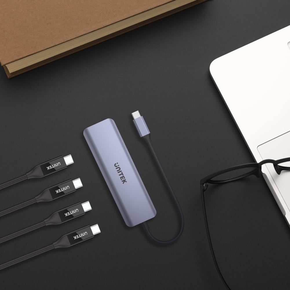 Ein Unitek USB-C-Hub, Kabel, Laptop, Brille und Buch auf dunkler Oberfläche.