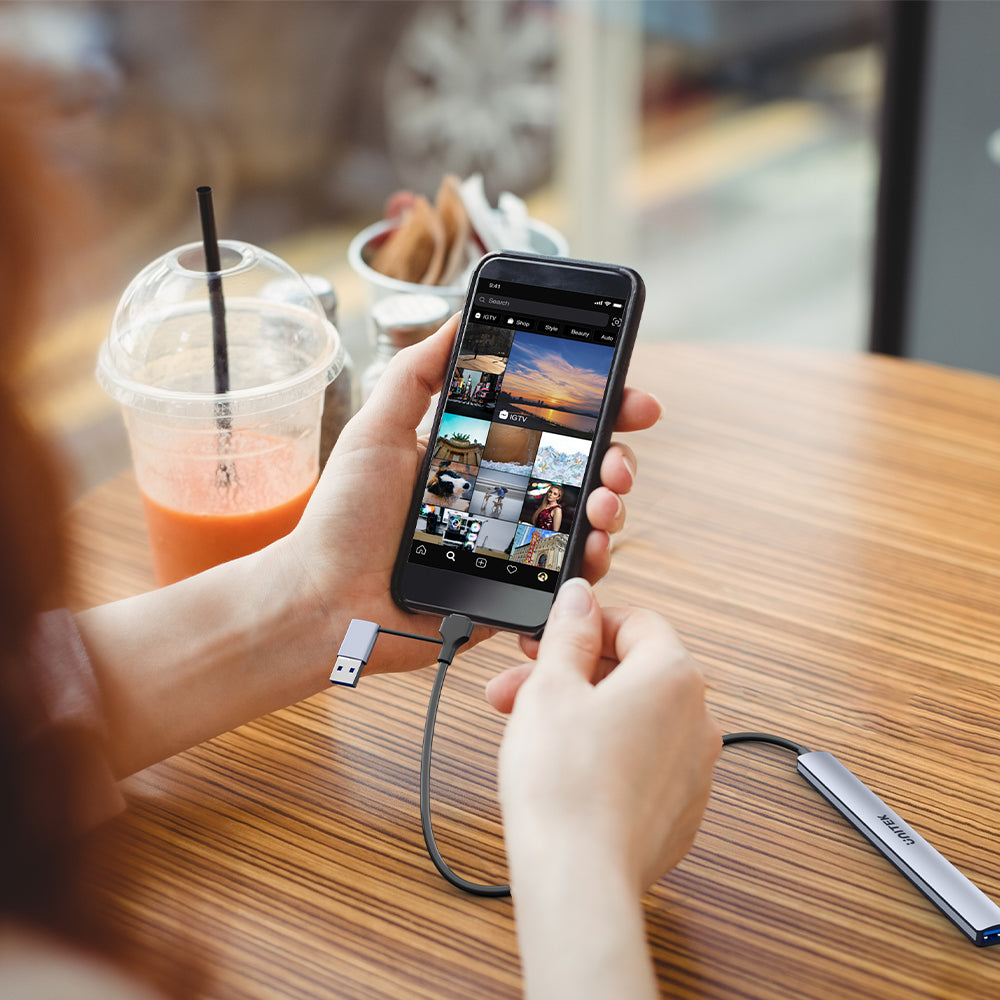 Una mano tiene uno smartphone collegato a un hub USB-C su un tavolo di legno.