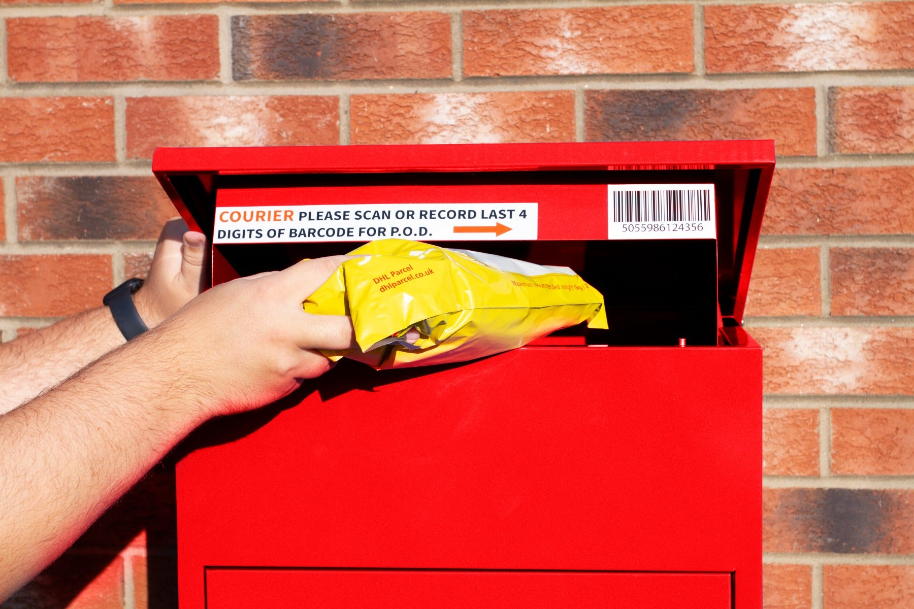 Una persona coloca un paquete amarillo en un buzón rojo frente a una pared de ladrillos.