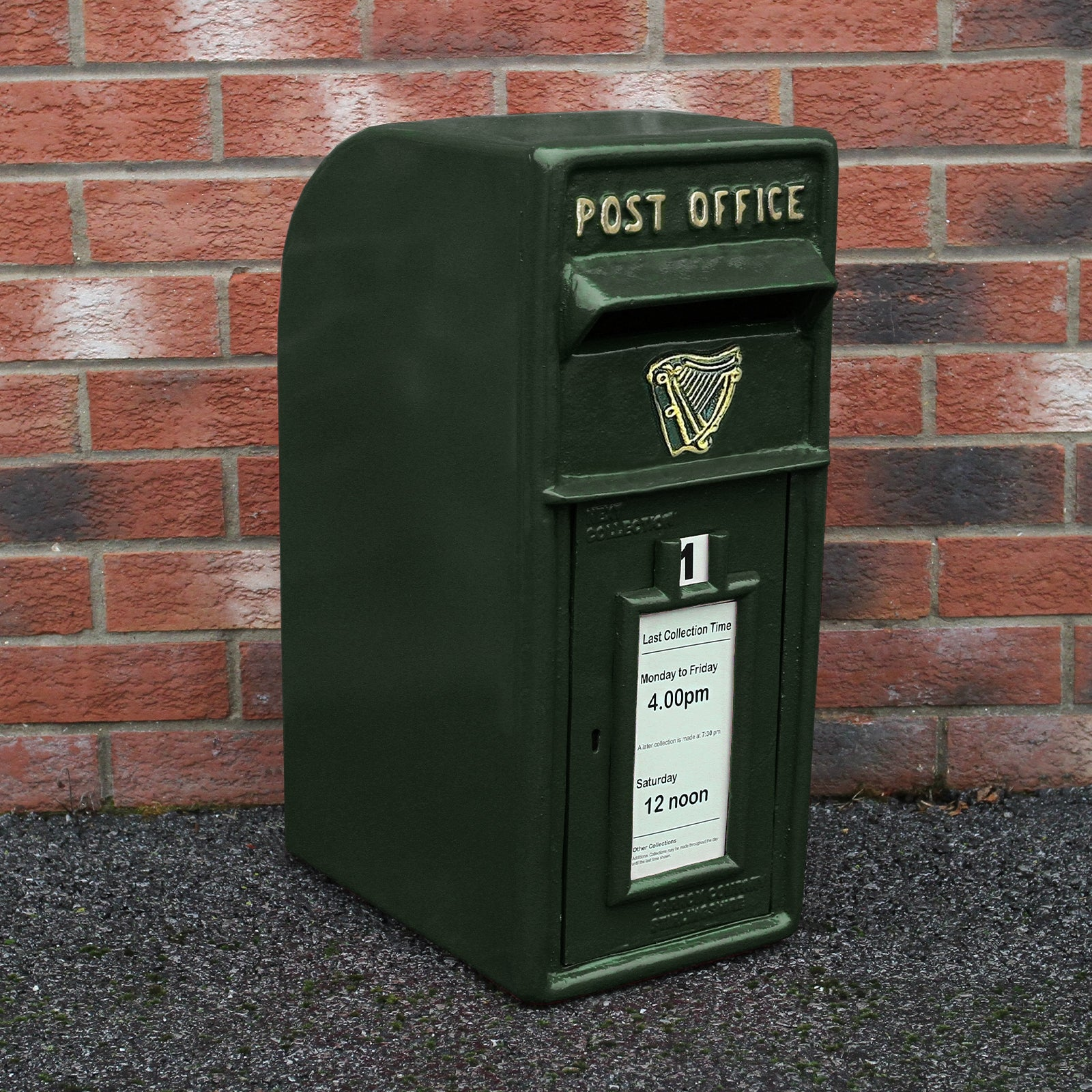 Un buzón verde oscuro con "Post Office" y emblema de arpa contra una pared de ladrillos.