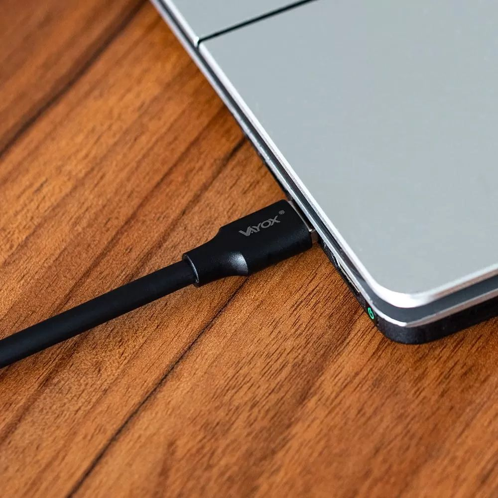 Ein schwarzes Vayox-Ladekabel, das in einen silbernen Laptop auf einem Holztisch gesteckt ist.