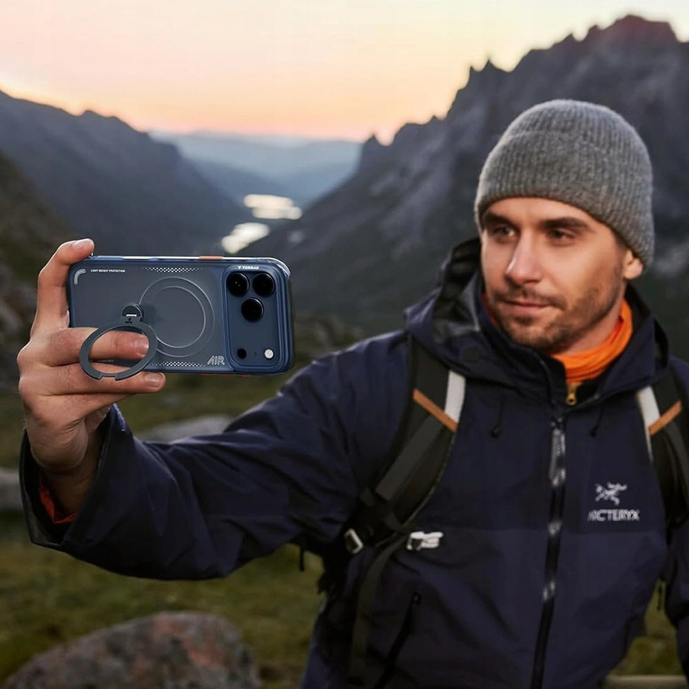 Bereli bir adam gün batımında dağ manzarasında akıllı telefonla selfie çekiyor.