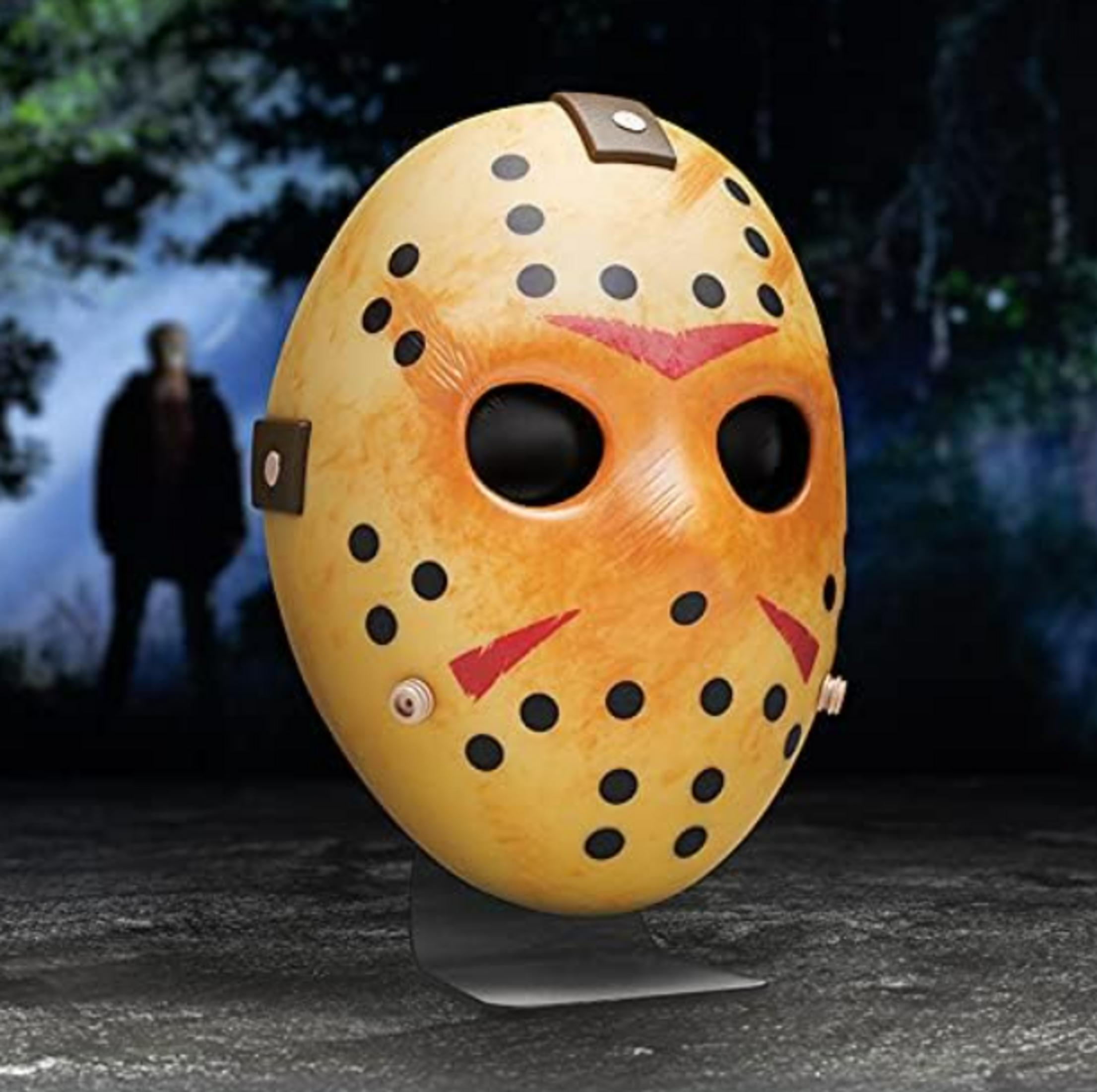 Eine gelbe Hockeymaske mit schwarzen Punkten und roten Markierungen, auf einem Ständer. Eine verschwommene Figur steht dahinter.