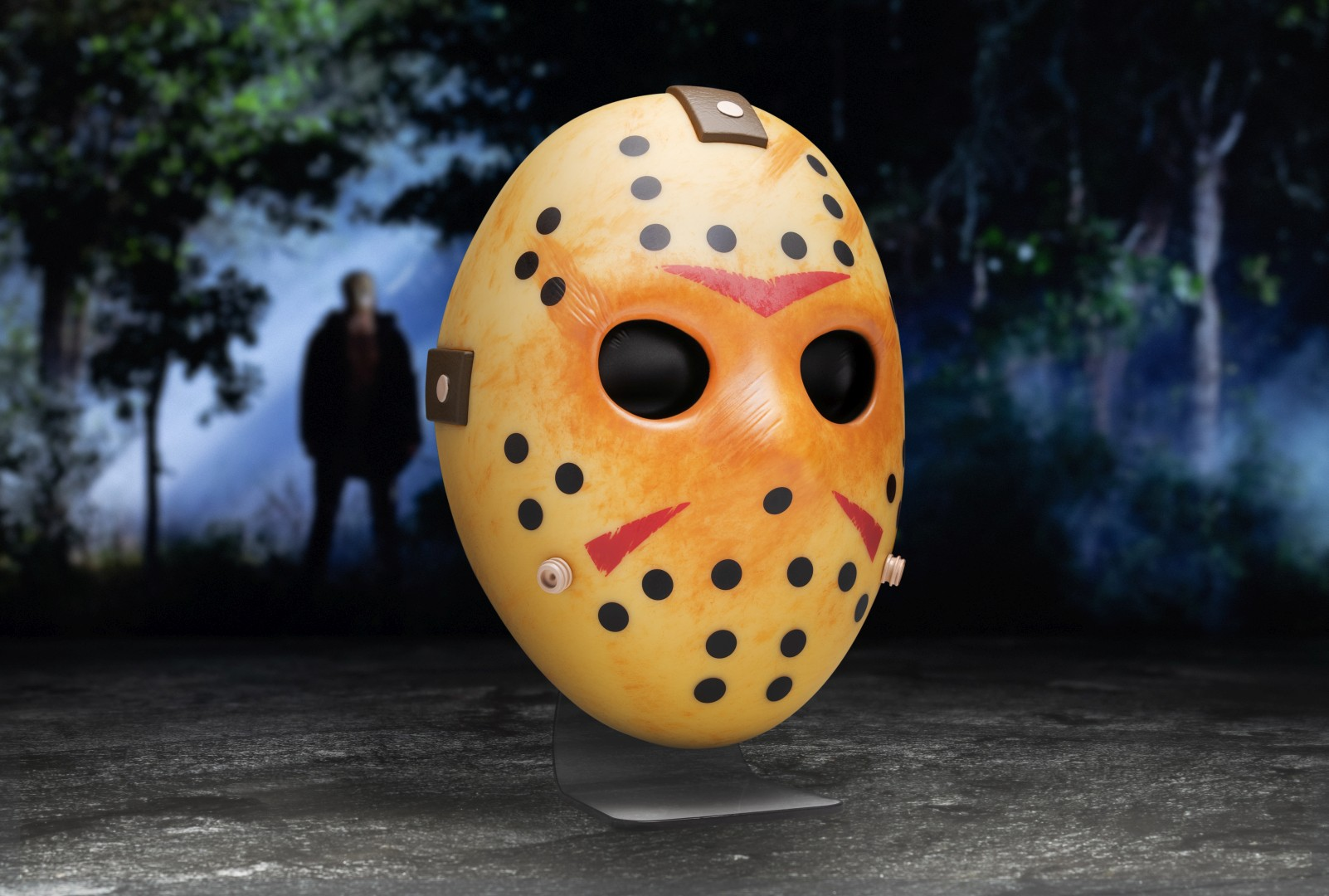 Gelbe Hockeymaske mit schwarzen Löchern und roten Dreiecken. Eine Figur steht im Hintergrund im Wald.
