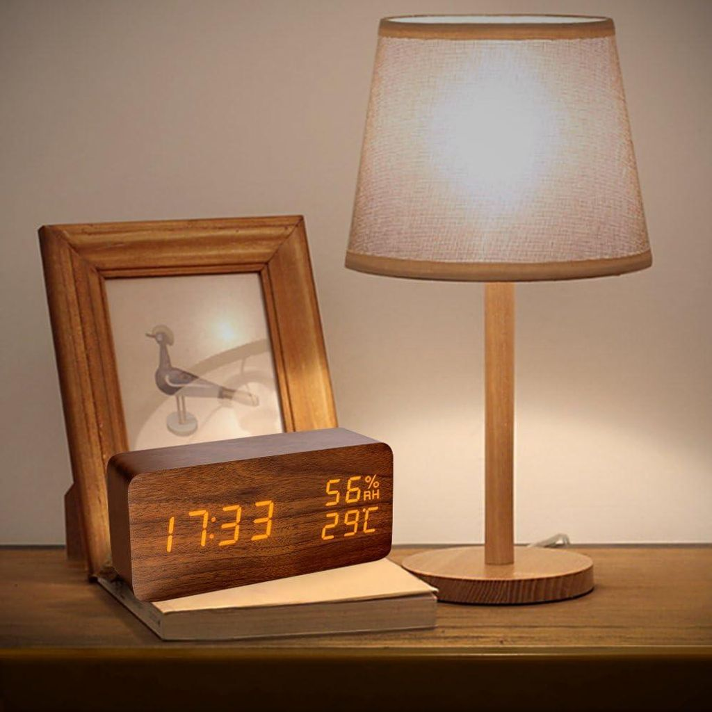 Holzuhr mit Digitalanzeige, Bilderrahmen mit Vogelmotiv und Lampe auf einem Nachttisch.
