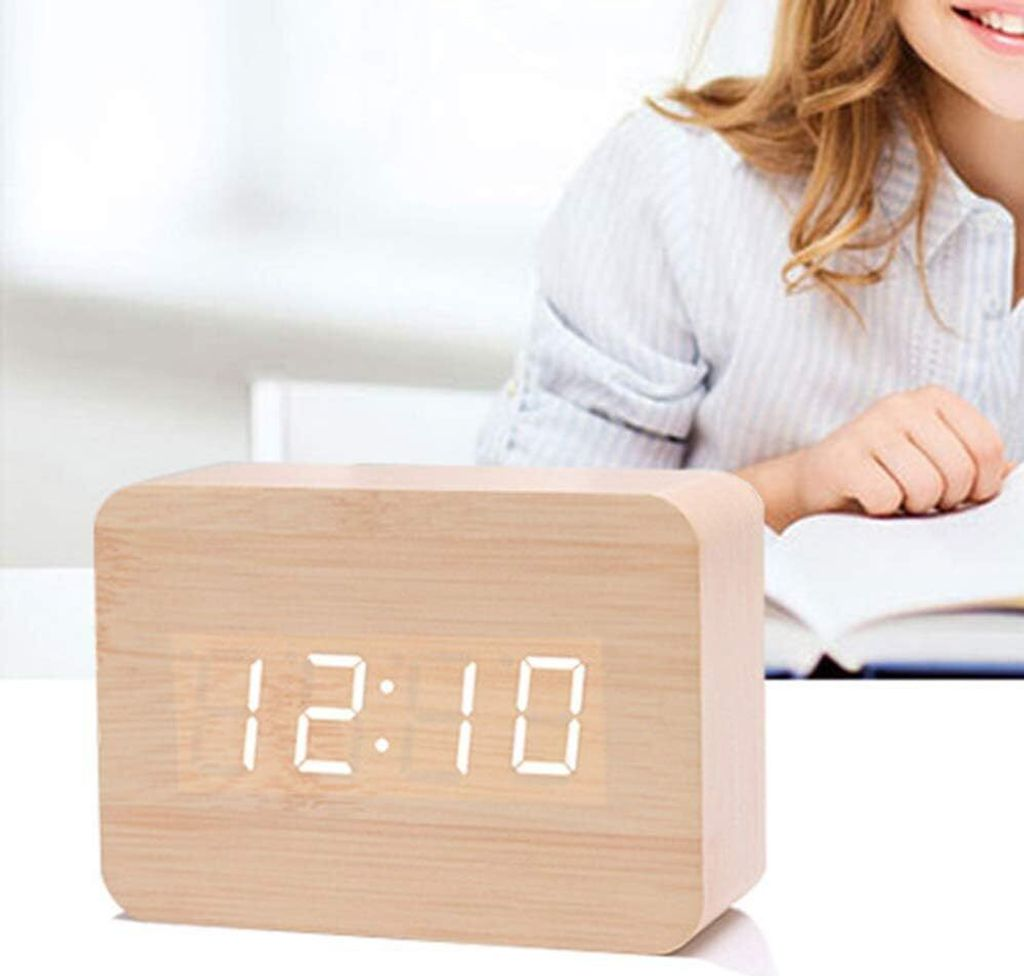 Ein hölzerner Digitalwecker zeigt 12:10 an, eine Person liest im Hintergrund.