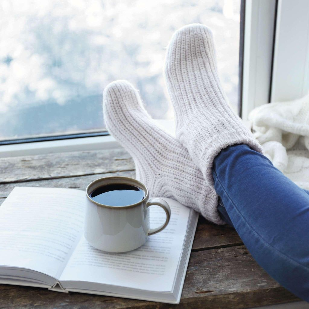 Person mit dicken Socken und Jeans liest Buch mit Kaffee am Fenster.