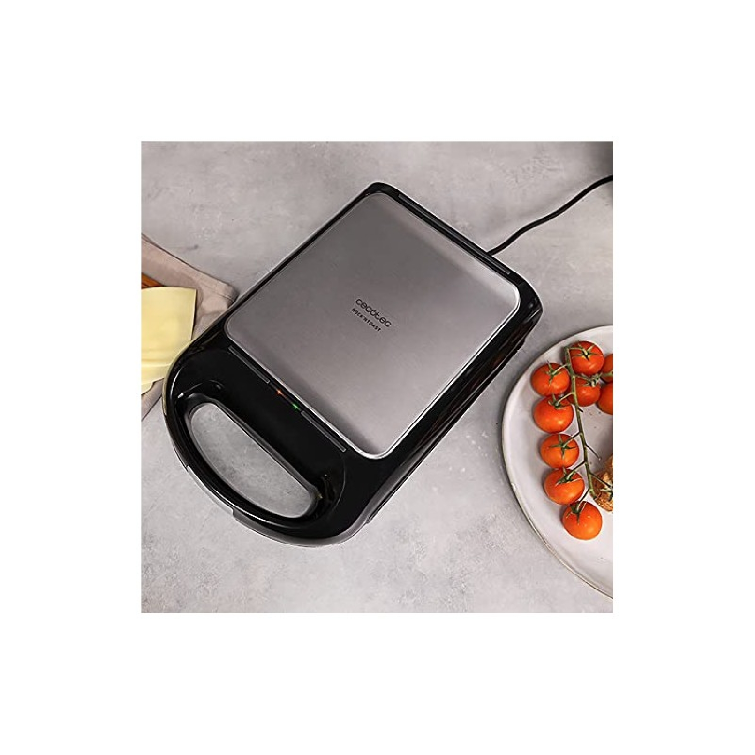 Una sandwichera cerrada con tapa de acero inoxidable junto a tomates.