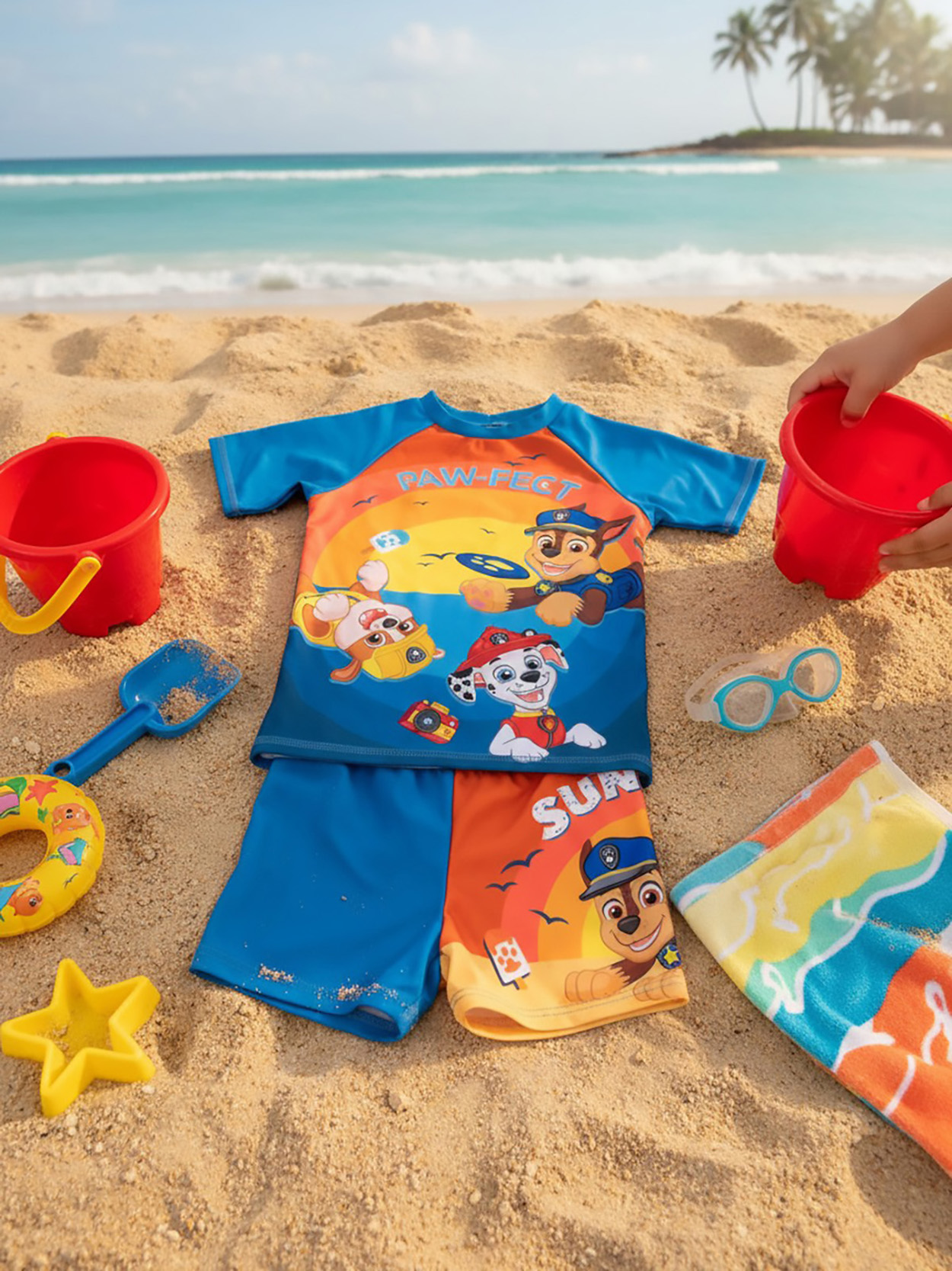 Paw Patrol-Badeanzug und Shorts am Sandstrand mit Eimern, Schaufel, Brille, Handtuch.