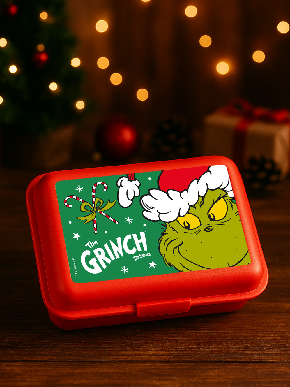 Rote Brotdose mit Grinch-Weihnachtsmotiv. Im Hintergrund Weihnachtsbaum und Lichter.