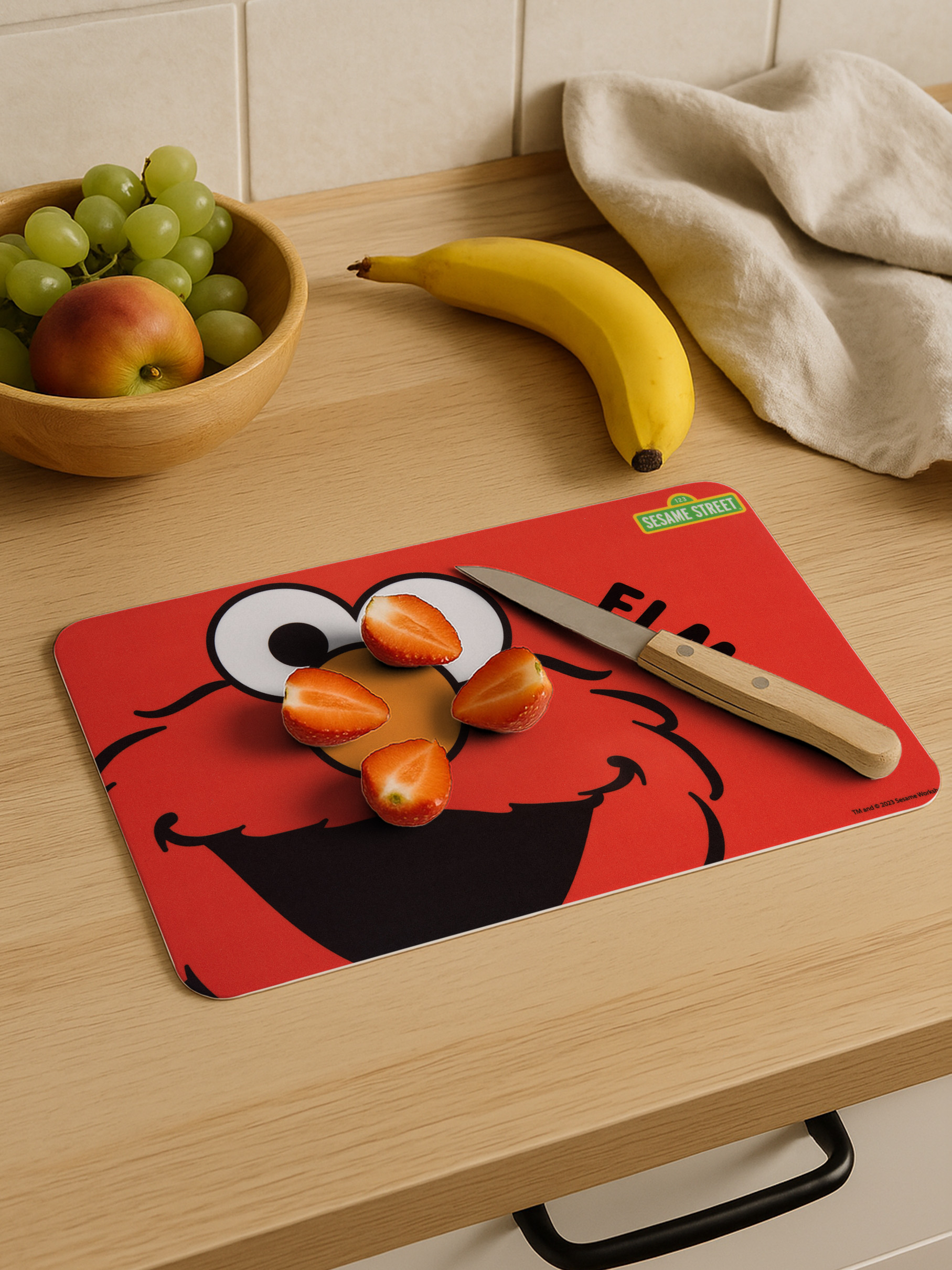 Schneidebrett mit Elmo-Motiv, Obst und Messer auf Holztisch.