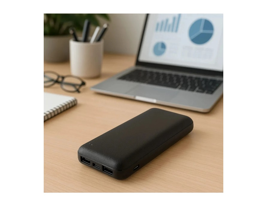 Siyah bir powerbank, dizüstü bilgisayar, gözlük ve not defterinin yanında ahşap bir masanın üzerinde duruyor.