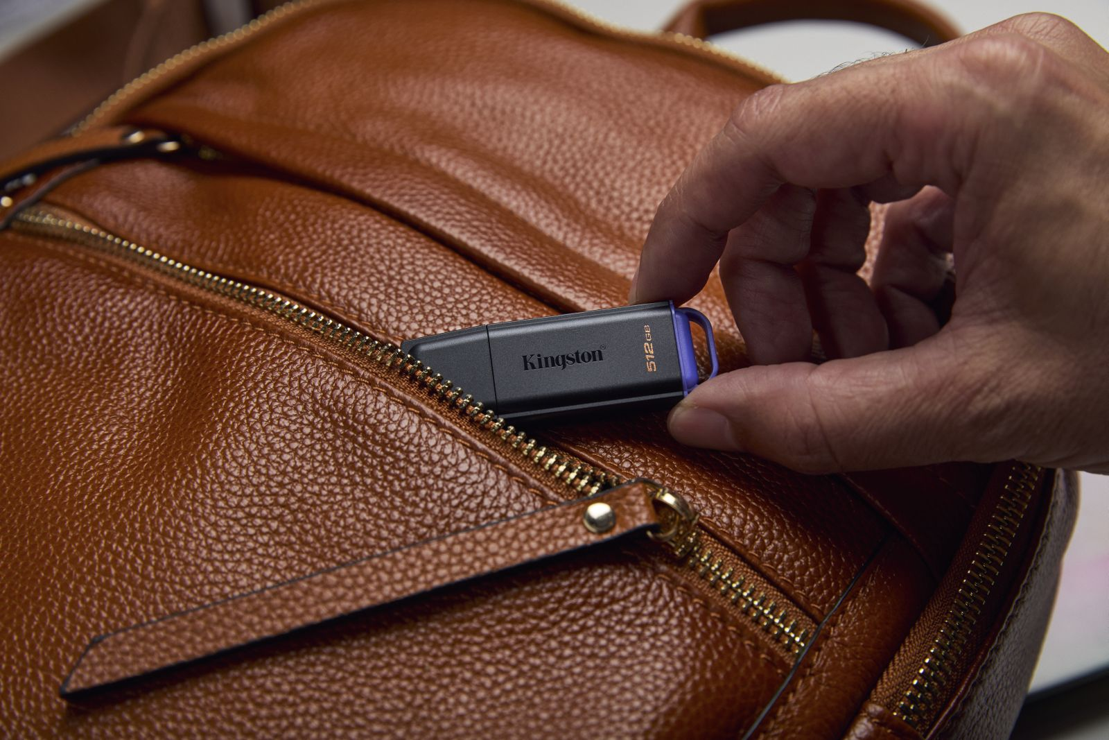 Dłoń trzyma czarny pendrive Kingston 512 GB na brązowej skórzanej torbie.