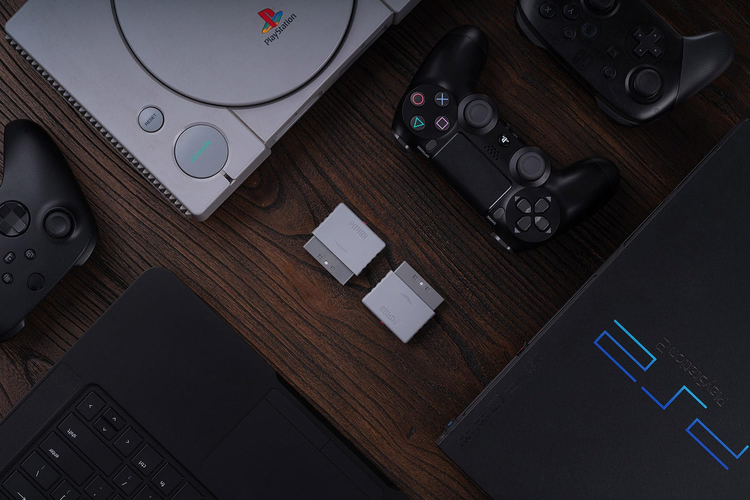 PlayStation 1, PS2, kontrolery i laptop na drewnie. Z przodu dwa adaptery.