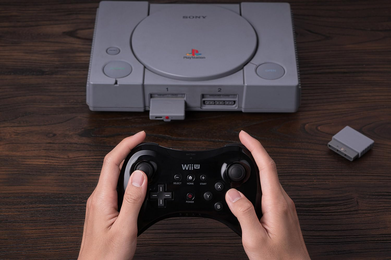 Dłonie trzymają czarny kontroler Wii U przed szarą konsolą PlayStation 1.