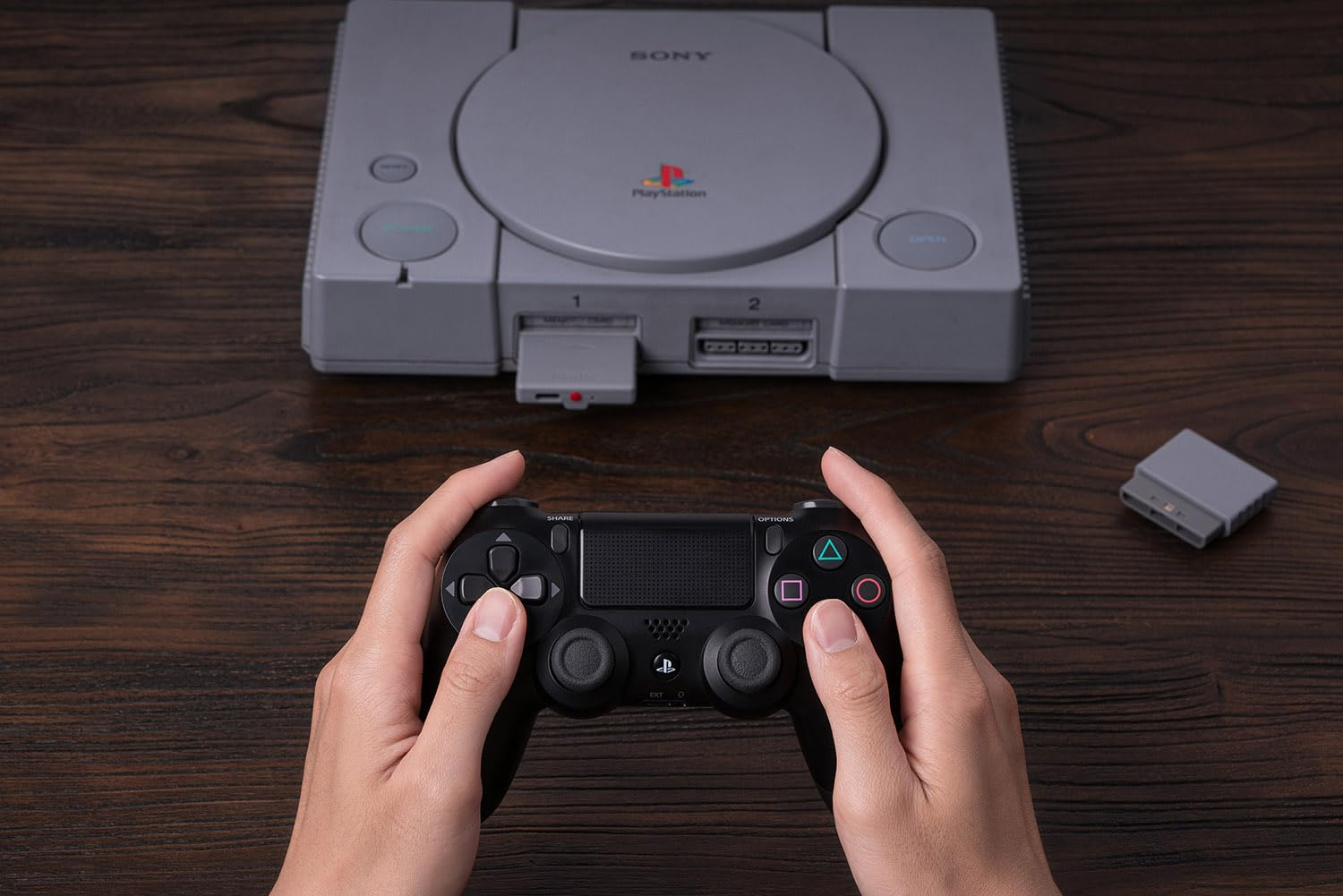 Dłonie trzymają czarny kontroler PlayStation przed szarą konsolą PlayStation.