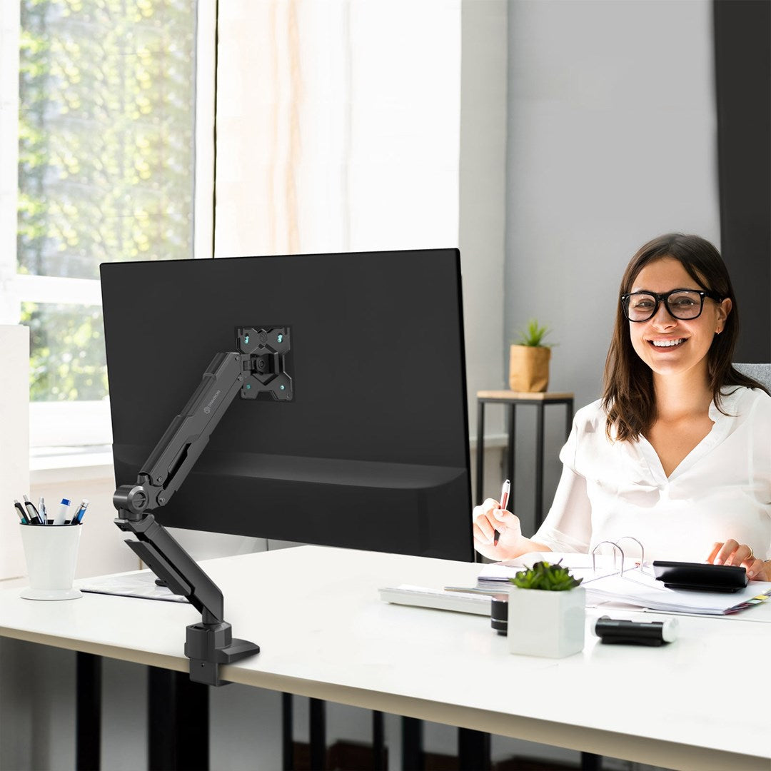 Kobieta siedzi przy biurku, uśmiecha się i patrzy do przodu. Monitor na ramieniu stoi przed nią.