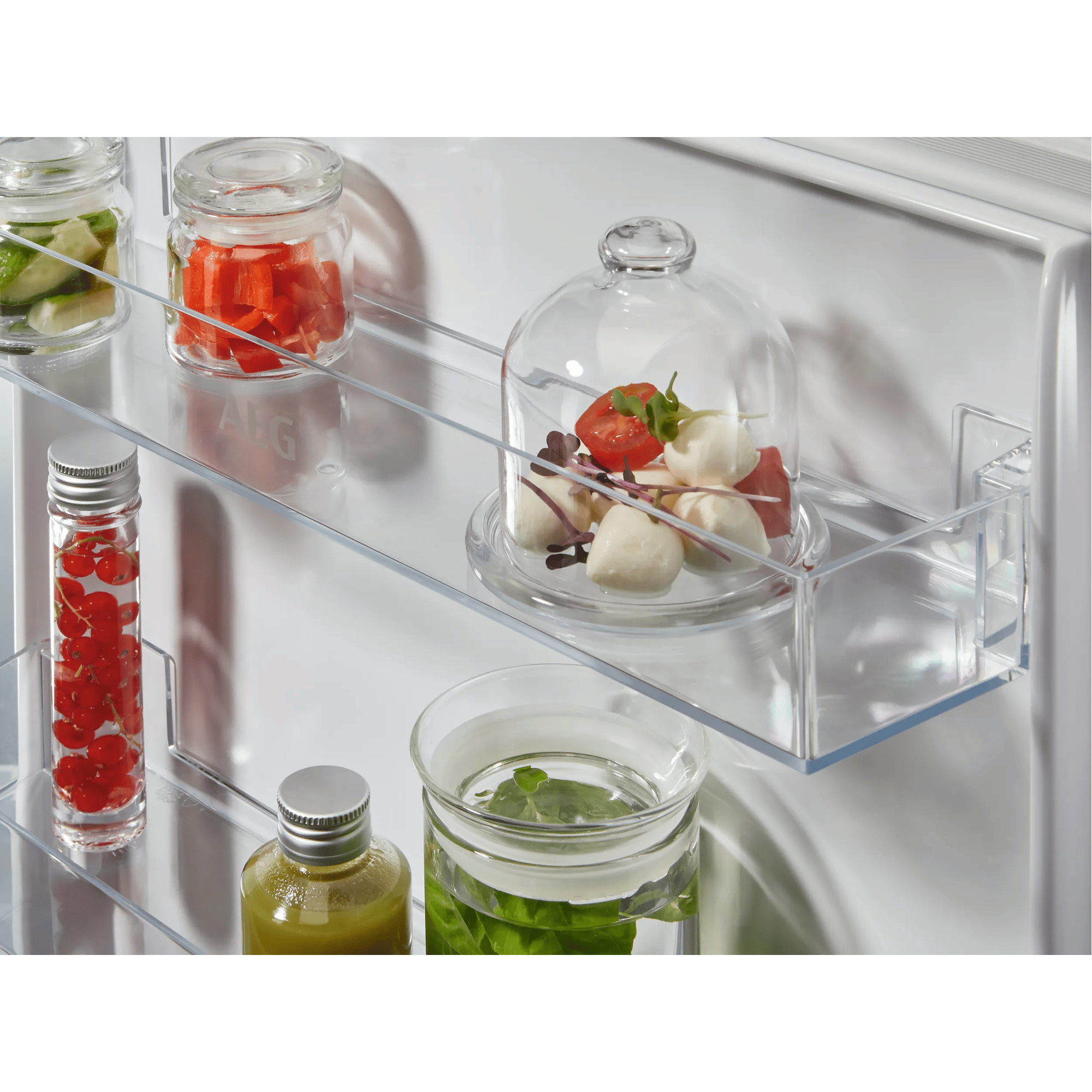 Estante de refrigerador con frascos: pepinos, pimientos rojos, mozzarella con tomates, grosellas rojas, zumo, agua de hierbas.
