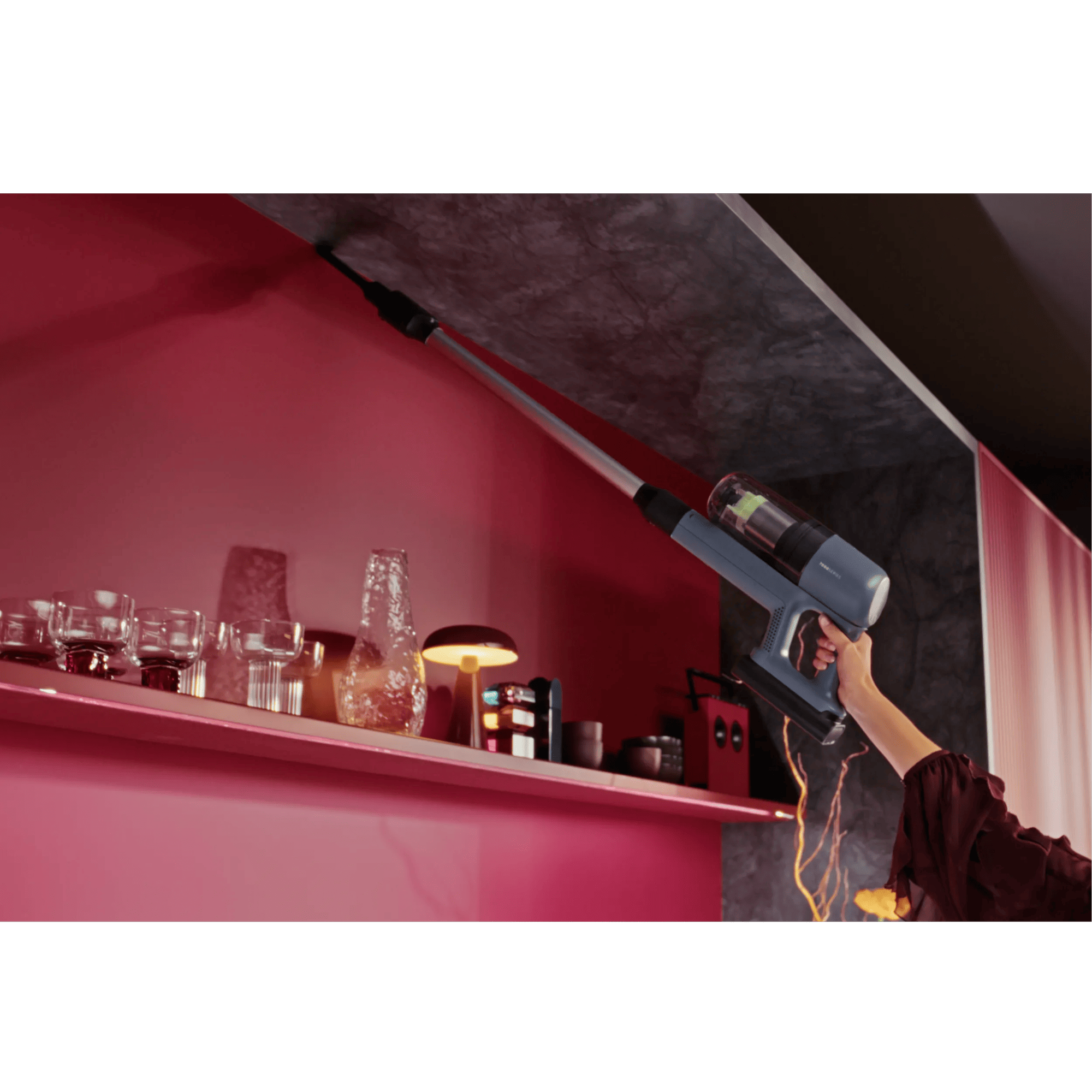 A hand holds a blue cordless vacuum cleaner being used to clean the ceiling.